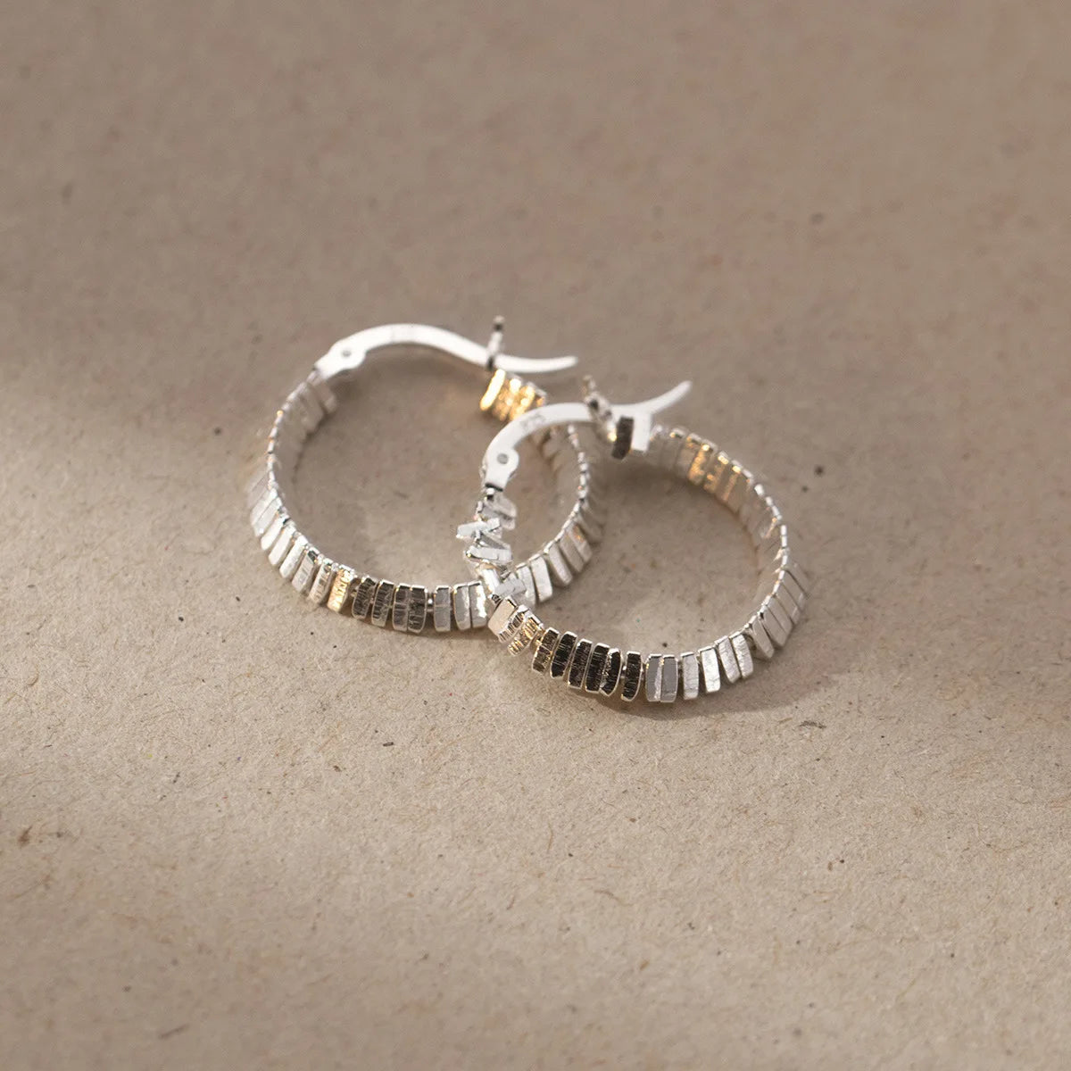  Zwei strukturierte Kreolen-Ohrringe (925er Silber) liegen auf einer hellbraunen, strukturierten Oberfläche.