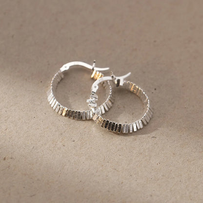  Zwei strukturierte Kreolen-Ohrringe (925er Silber) liegen auf einer hellbraunen, strukturierten Oberfläche.