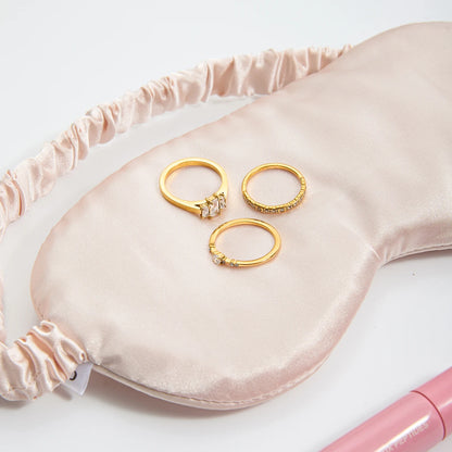 Drei glänzende goldene Ringe mit Zirkoniasteinen, elegant drapiert auf einer cremefarbenen oder hellrosa Satin-Schlafmaske.