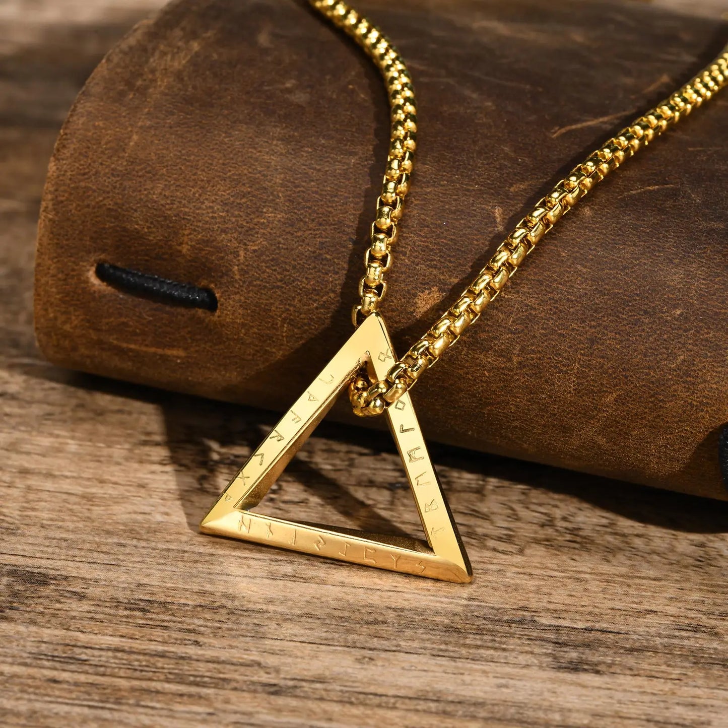 Detailansicht einer goldfarbenen Edelstahlkette mit offenem dreieckigen Anhänger, liegend auf einer braunen Lederoberfläche und Holzhintergrund.
