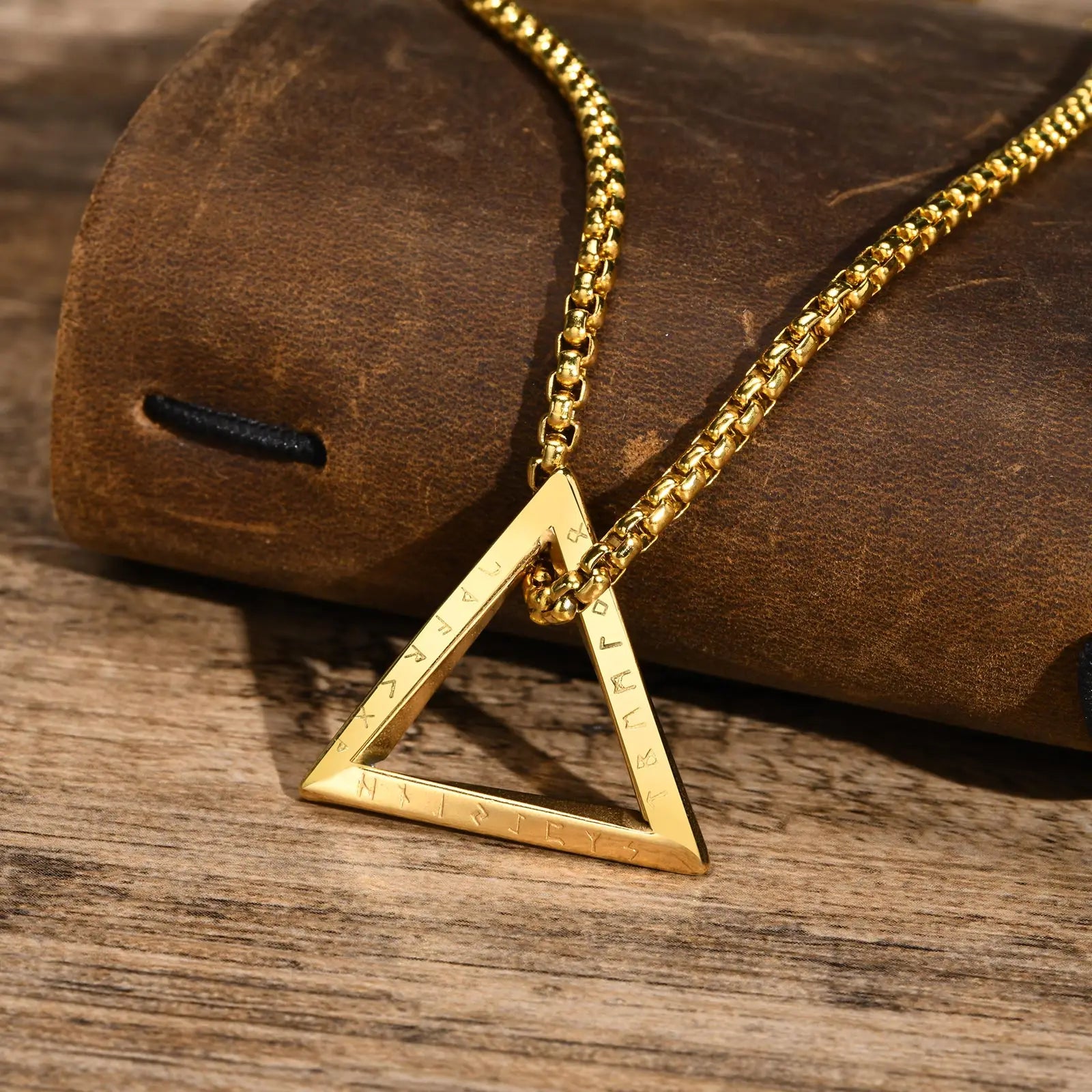 Detailansicht einer goldfarbenen Edelstahlkette mit offenem dreieckigen Anhänger, liegend auf einer braunen Lederoberfläche und Holzhintergrund.
