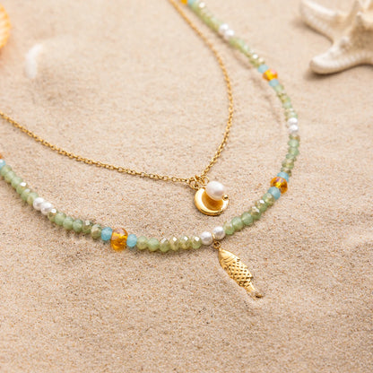 Maritime Halsketten im Sand: 18K vergoldete Edelstahlkette mit Süßwasserperle und Plättchen neben einer Perlenkette in Hellgrün und Weiß mit einem goldenen Fisch-Anhänger, dekoriert mit Seestern.