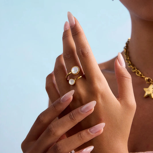 Nahaufnahme einer Hand mit gepflegten Fingernägeln, die einen offenen Wickelring aus 18K vergoldetem Edelstahl mit zwei runden, weiß schimmernden Elementen trägt.