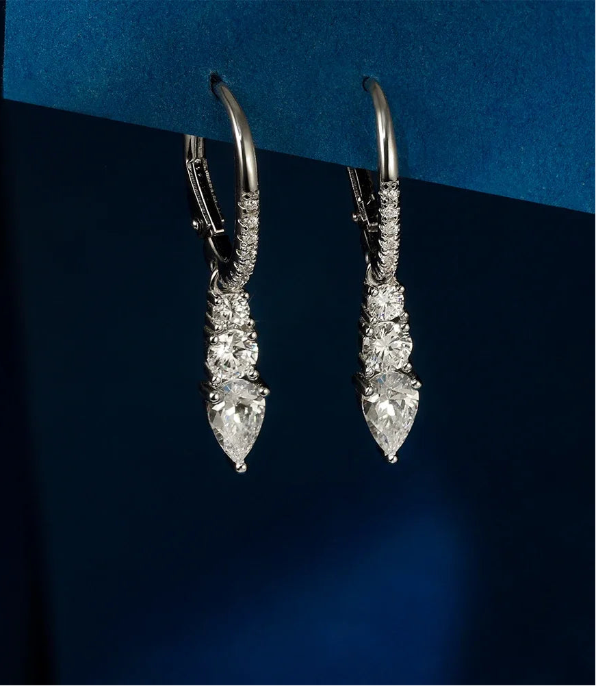 Freigestelltes Paar hängender Ohrringe aus reinem 925er Sterling Silber, jeweils besetzt mit drei gestuften, tropfenförmigen Zirkonia-Steinen und einem Zirkonia-besetzten Kreolenbügel. Das 'Real 925 Sterling Silver' Siegel ist unten rechts erkennbar.