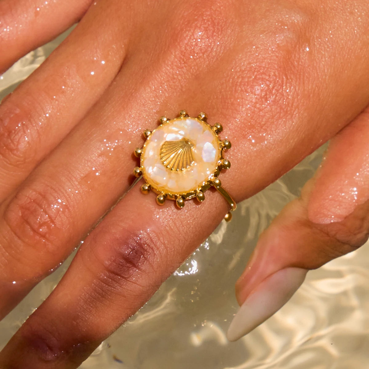 Hand einer Person, die einen ovalen Ring mit beigefarbenem Stein und Muschel-Symbol aus 18K vergoldetem Edelstahl über dem Wasser hält.