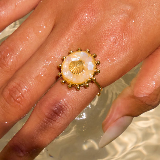 Hand einer Person, die einen ovalen Ring mit beigefarbenem Stein und Muschel-Symbol aus 18K vergoldetem Edelstahl über dem Wasser hält.