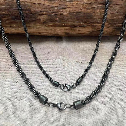 Zwei schwarze Edelstahl-Kordelketten in unterschiedlichen Längen (Lagen-Look) mit Karabinerverschlüssen, hängend vor einem hellen Hintergrund.