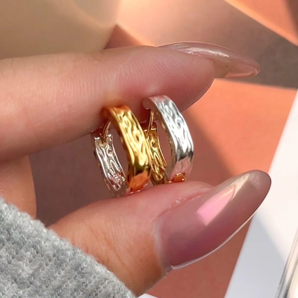 Nahaufnahme der gehämmerten Creolen in Silber und Gold, gehalten zwischen zwei Fingerspitzen.