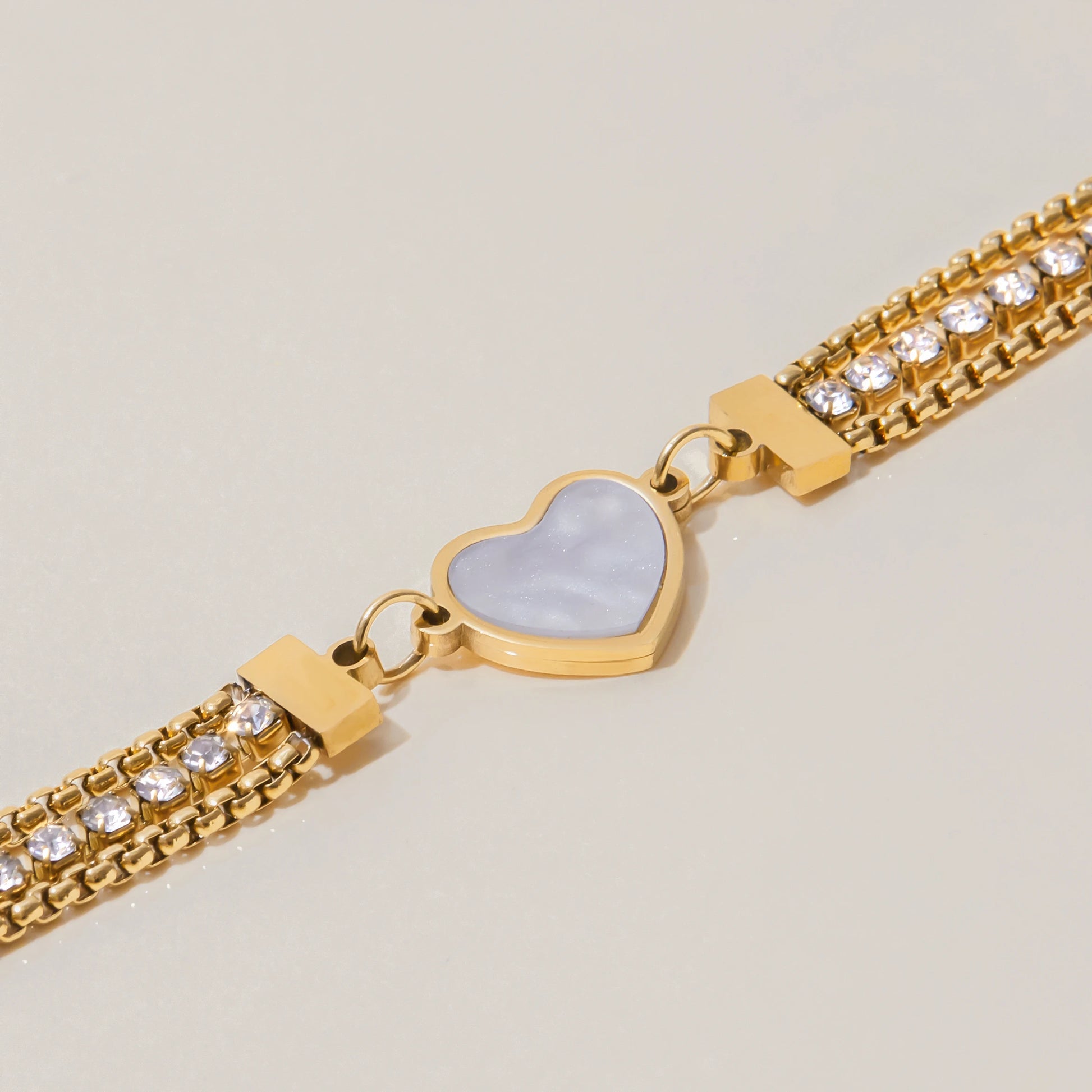 Detailaufnahme eines 18K vergoldeten Edelstahl-Armbands: Fokus auf das eingefasste Herz-Element in weißer Perlmutt-Optik und die angrenzenden Zirkonia-Strassketten.