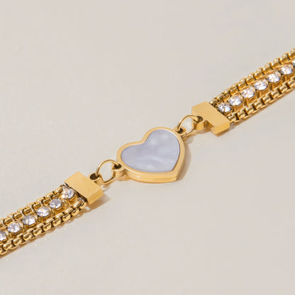 Detailaufnahme eines 18K vergoldeten Edelstahl-Armbands: Fokus auf das eingefasste Herz-Element in weißer Perlmutt-Optik und die angrenzenden Zirkonia-Strassketten.