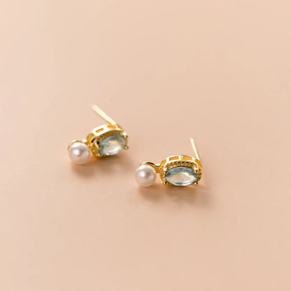 Ein Paar elegante Ohrringe aus 18K vergoldetem 925er Silber mit hellblauem Schmuckstein und kleiner Perle, flach arrangiert auf beigefarbenem Hintergrund.
