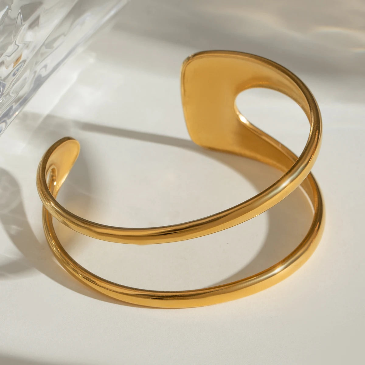 Breite goldene Armspange aus Edelstahl mit glänzendem Finish, im hellen Sonnenlicht mit markanten Schatten fotografiert.