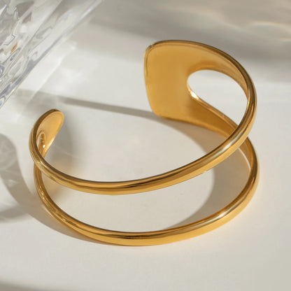 Breite goldene Armspange aus Edelstahl mit glänzendem Finish, im hellen Sonnenlicht mit markanten Schatten fotografiert.