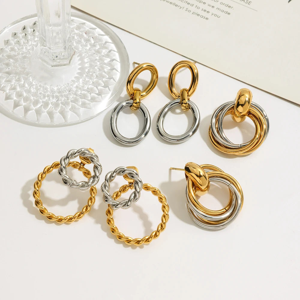 Verschiedene goldene und silberne Ohrringe aus Edelstahl mit 18K PVD-Vergoldung und PVD-Beschichtung. Die Ohrringe haben verschlungene oder ineinandergreifende Ringdesigns, die in drei verschiedenen Variationen auf einer hellen Oberfläche liegen.