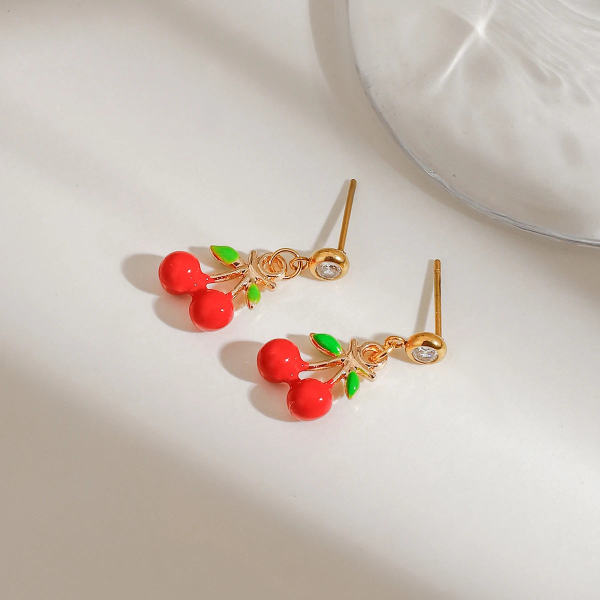Detailaufnahme von zwei 18K vergoldeten Edelstahl-Ohrsteckern mit hängendem Kirschen-Motiv in leuchtendem Rot und Grün.