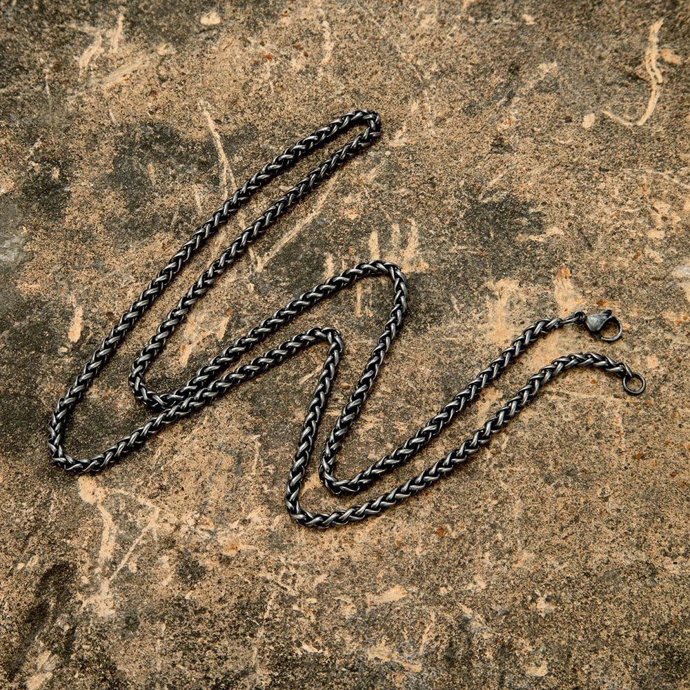 Nahaufnahme der schwarzen, geflochtenen Edelstahl-Kette (Weizenmuster) im Vintage-Stil. Die Kette liegt zusammengerollt auf einem dunklen, steinernen Untergrund.