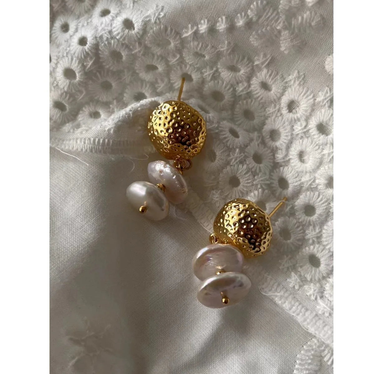 Nahaufnahme eines Paares goldener Ohrstecker mit gehämmertem Metallteil und zwei flachen, unregelmäßigen Baroque-Süßwasserperlen, die auf weißer Lochspitze liegen.