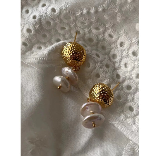 Nahaufnahme eines Paares goldener Ohrstecker mit gehämmertem Metallteil und zwei flachen, unregelmäßigen Baroque-Süßwasserperlen, die auf weißer Lochspitze liegen.