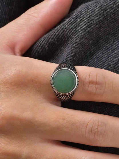 Nahaufnahme des gemusterten Edelstahlrings mit grünem Oval-Stein, getragen am Ringfinger vor grauem Denim-Stoff.