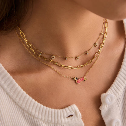 Eine Frau trägt drei verschiedene Halsketten im Layering-Stil, wobei die unterste Kette die goldvergoldete Edelstahlkette mit dem rosa Dackel-Motiv ist.