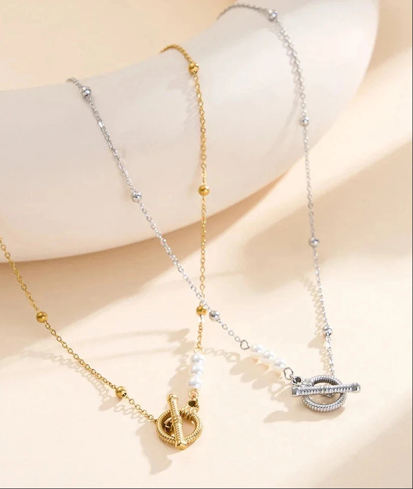 Zwei Edelstahl-Halsketten mit Knebelverschluss und Muschelperlen, ein Modell in Gold und eines in Silber, drapiert auf einem hellen Podest.