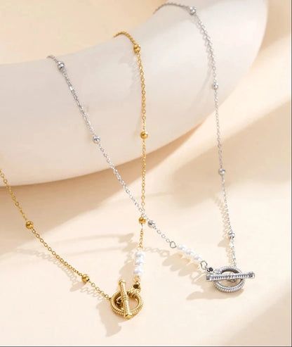 Zwei Edelstahl-Halsketten mit Knebelverschluss und Muschelperlen, ein Modell in Gold und eines in Silber, drapiert auf einem hellen Podest.