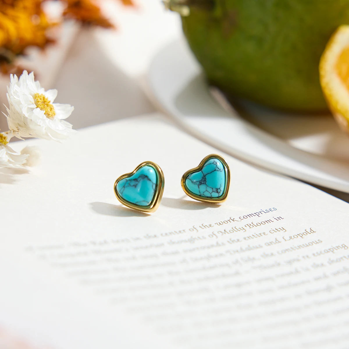 Paar Herz-Ohrstecker aus 18K vergoldetem Edelstahl mit blauem Türkisstein auf einem Tisch, im Hintergrund eine Avocado.