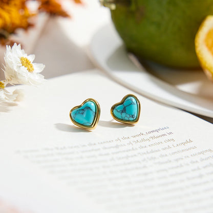 Paar Herz-Ohrstecker aus 18K vergoldetem Edelstahl mit blauem Türkisstein auf einem Tisch, im Hintergrund eine Avocado.