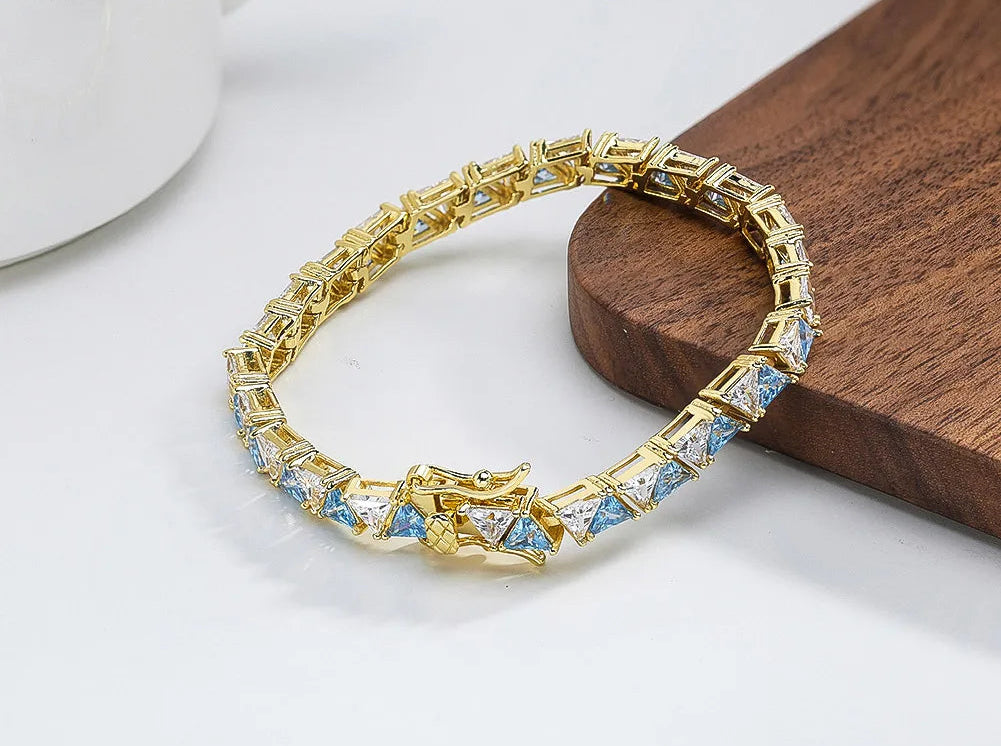Goldenes Armband aus 14K vergoldetem 925er Silber, besetzt mit funkelnden hellblauen AAAAA Zirkonia-Steinen, liegend auf einem hellen Schreibtisch mit unscharfem Hintergrund.