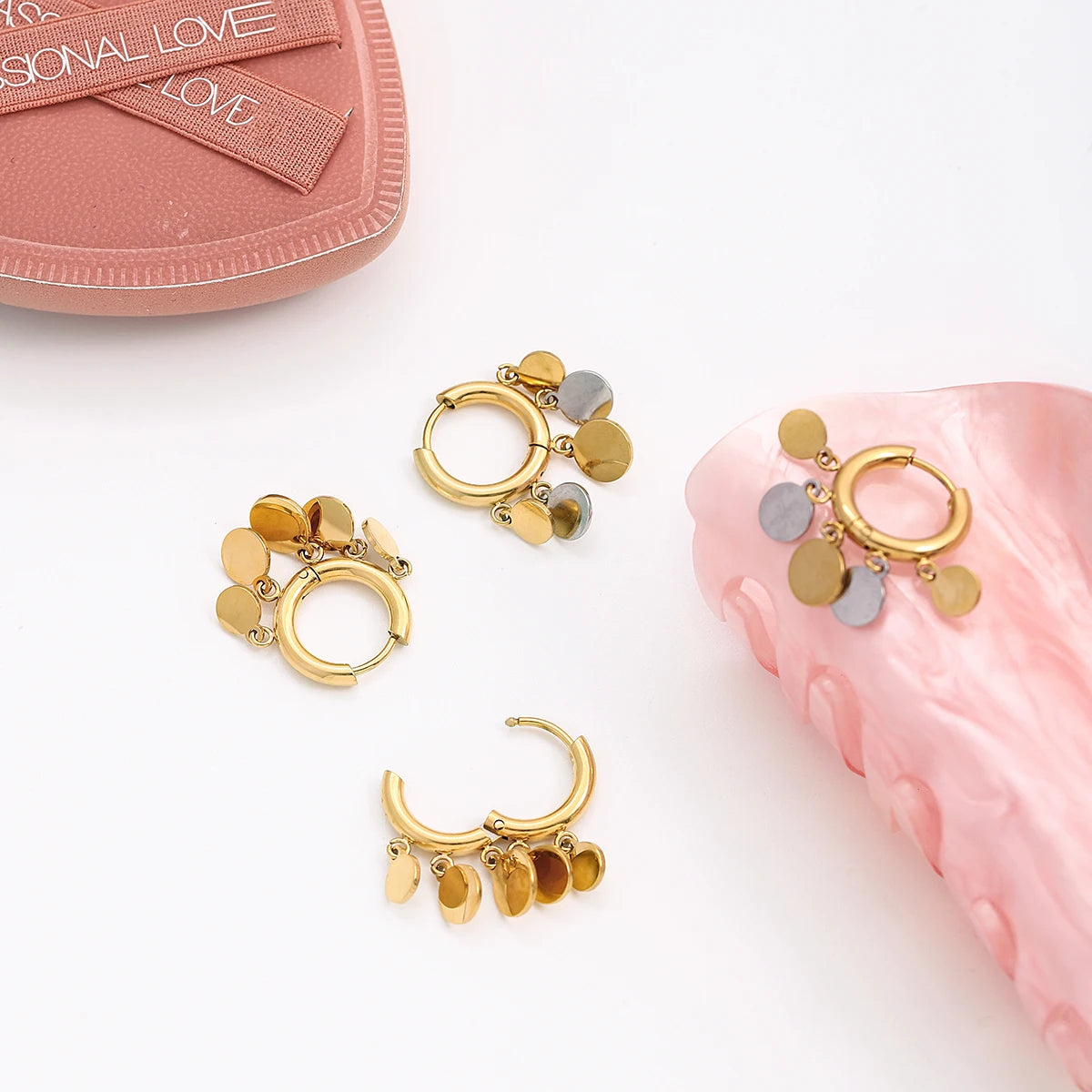 Stilvolle Produktfotografie der goldenen Plättchen-Creolen auf heller Oberfläche neben rosafarbenen Accessoires.