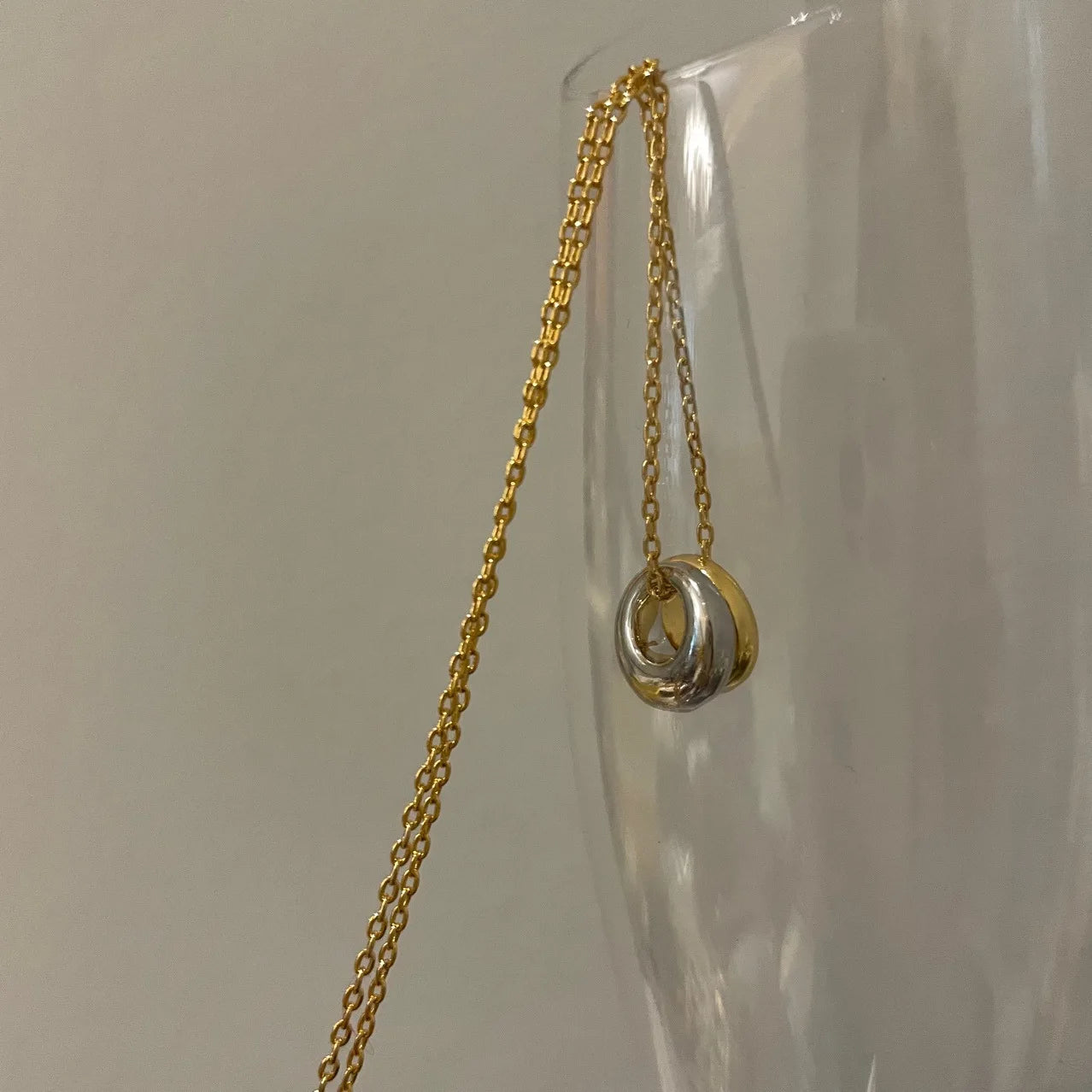 Feine goldene Ankerkette mit Bicolor-Anhängern in Gold und Silber, aufgehängt an einem Glas.