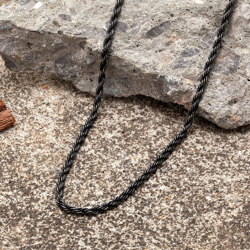  Ganze Ansicht der schwarzen Edelstahl-Kordelkette im Vintage-Stil auf einem rauen, steinernen Boden. Die Kette ist gewunden dargestellt.