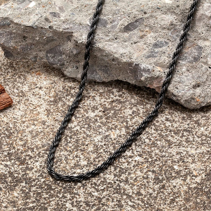  Ganze Ansicht der schwarzen Edelstahl-Kordelkette im Vintage-Stil auf einem rauen, steinernen Boden. Die Kette ist gewunden dargestellt.