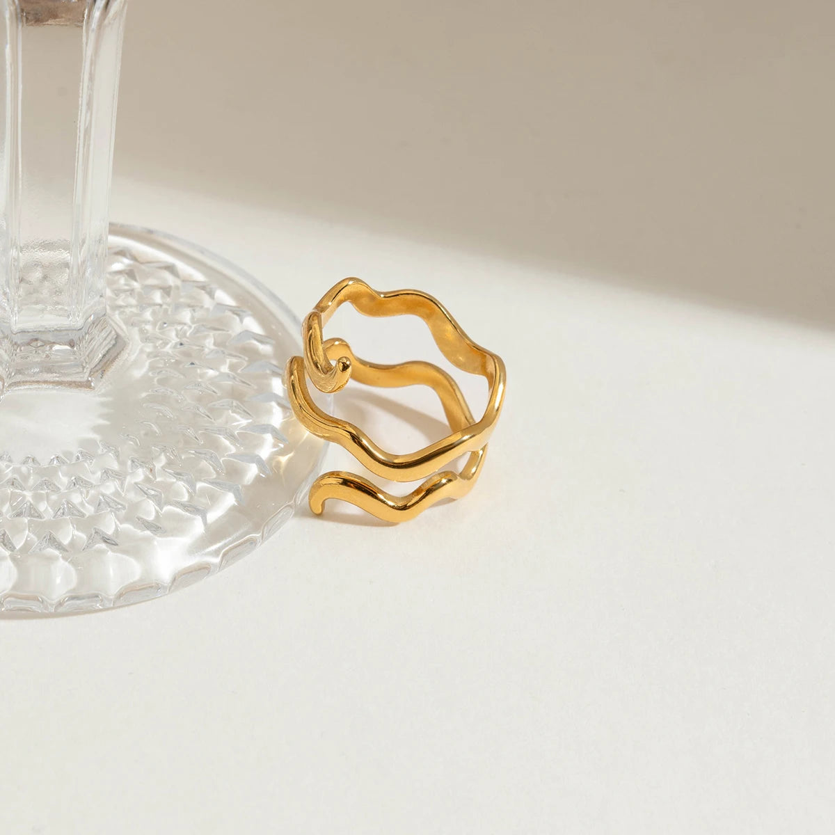 Elegantes Stillleben-Foto des gewellten, vergoldeten Edelstahlrings, platziert neben dem Fuß eines Kristallglases, um die glänzende Oberfläche hervorzuheben.