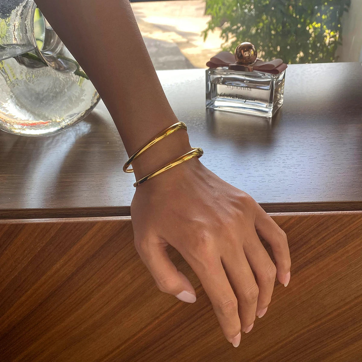 Nahaufnahme einer Hand, die einen modernen, zweireihigen Armreif aus 18K vergoldetem Edelstahl trägt, im Hintergrund steht ein Parfümflakon auf einer Holzoberfläche.
