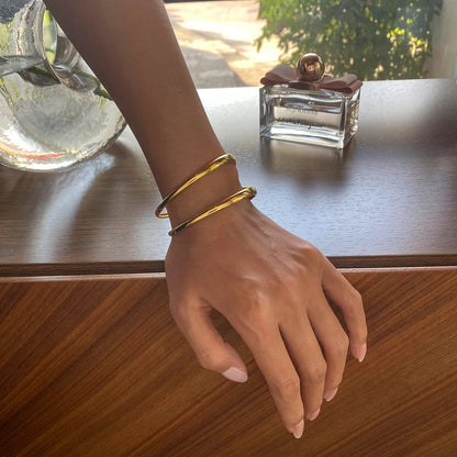 Nahaufnahme einer Hand, die einen modernen, zweireihigen Armreif aus 18K vergoldetem Edelstahl trägt, im Hintergrund steht ein Parfümflakon auf einer Holzoberfläche.