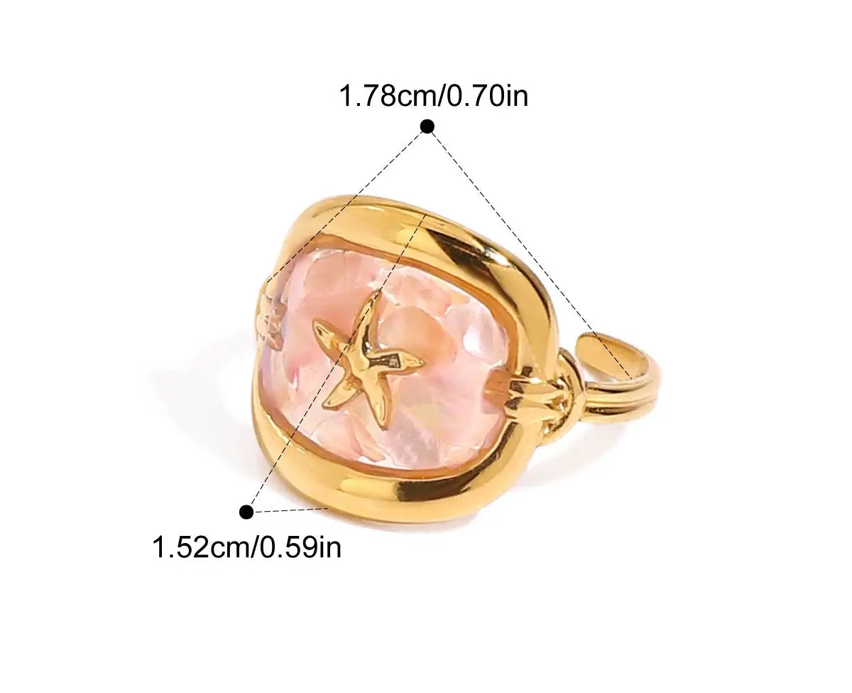 Nahaufnahme eines Rings aus 18K vergoldetem Edelstahl mit einem rosafarbenen Stein und einem erhabenen Muschel-Symbol. Der Ring hat eine Innendurchmesser von 1,78 cm und eine Breite von 1,52 cm.